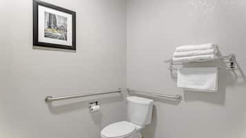 Ein Badezimmer mit einer weißen Toilette, Haltegriffen, einem gerahmten Bild an der Wand und einem Handtuchhalter mit gefalteten Handtüchern.