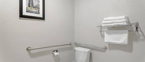 A bathroom with a white toilet, handrails, a framed picture on the wall, and a towel rack with folded towels.
