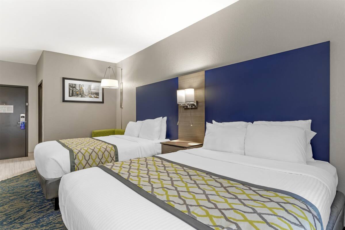 A hotel room with two beds, a desk, and a chair. There is a framed picture on the wall and a door with a lock.