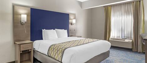A hotel room with a large bed, a bedside table, a nightstand, a wall-mounted lamp, an air conditioning unit, and a window with curtains.