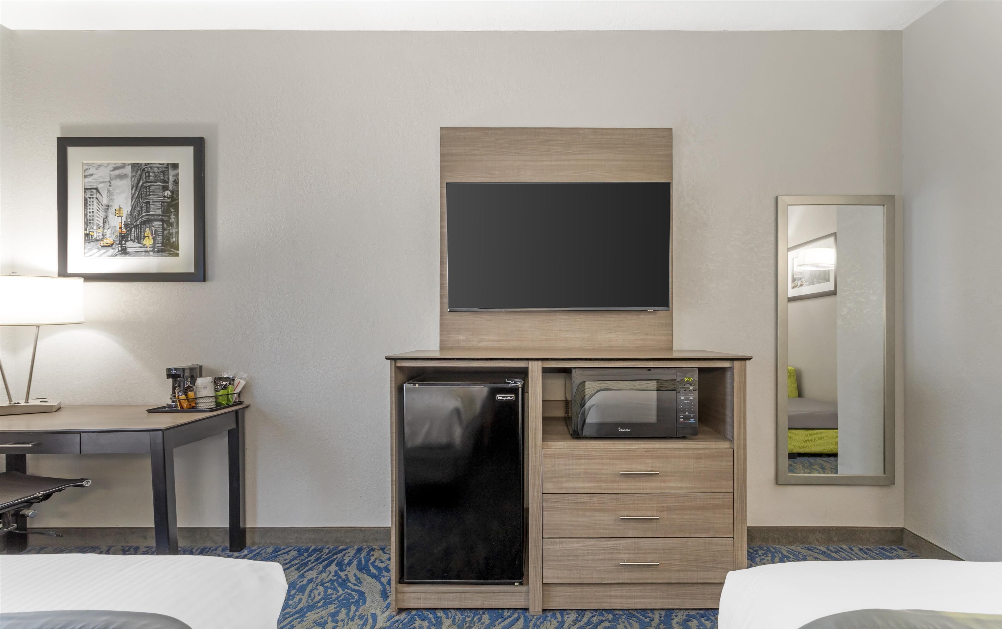 A hotel room with a bed, a desk with a lamp, a refrigerator, a microwave, a flat-screen TV, and a mirror.