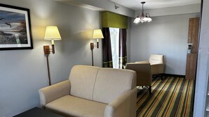 Hotellihuone, jossa on sohva, kaksi lamppua, tuoli, pöytä, raidallinen matto ja ovi, jossa on puupaneeli.