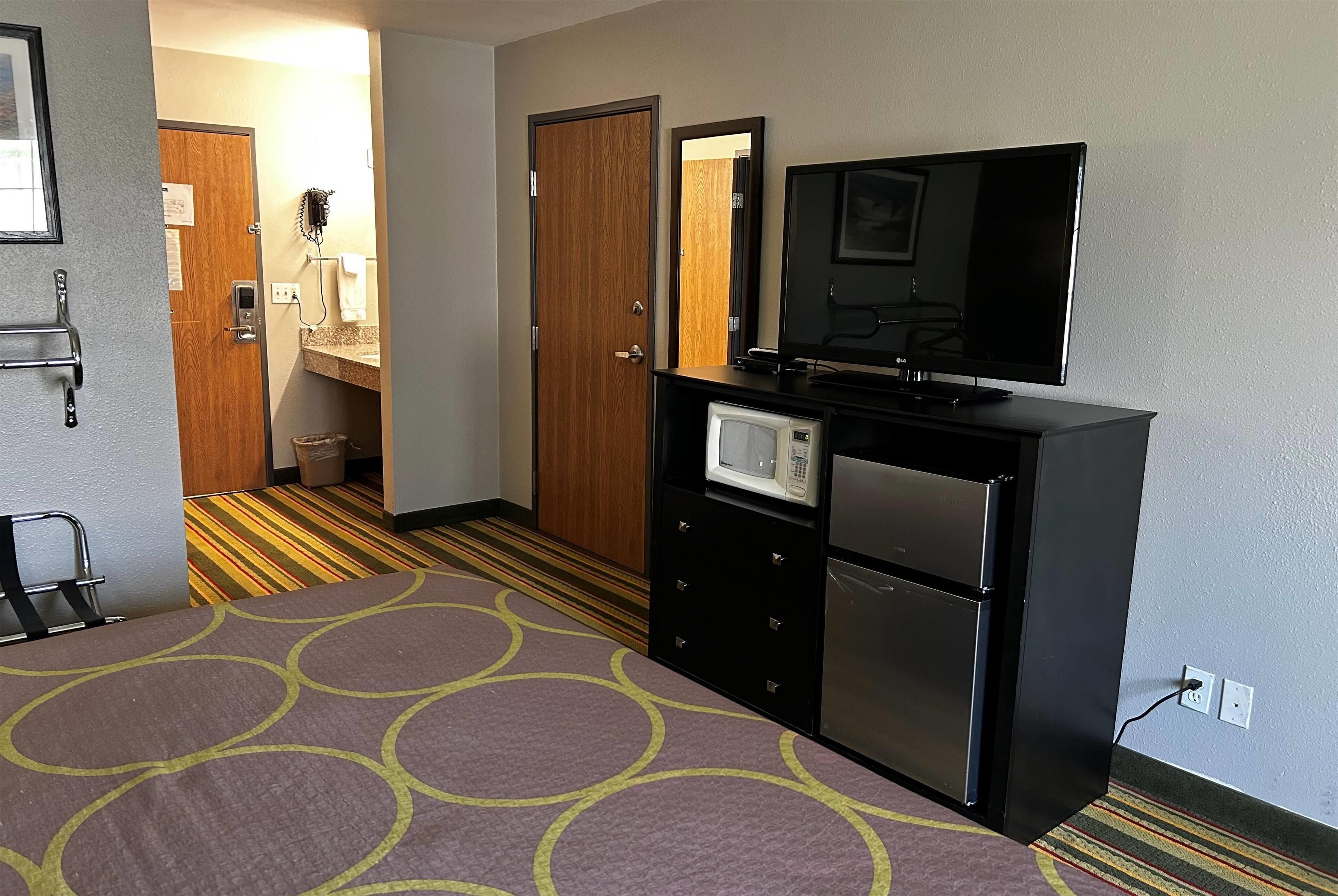 A hotel room with a bed, a TV, a microwave, a refrigerator, a desk, and a bathroom visible through an open door.