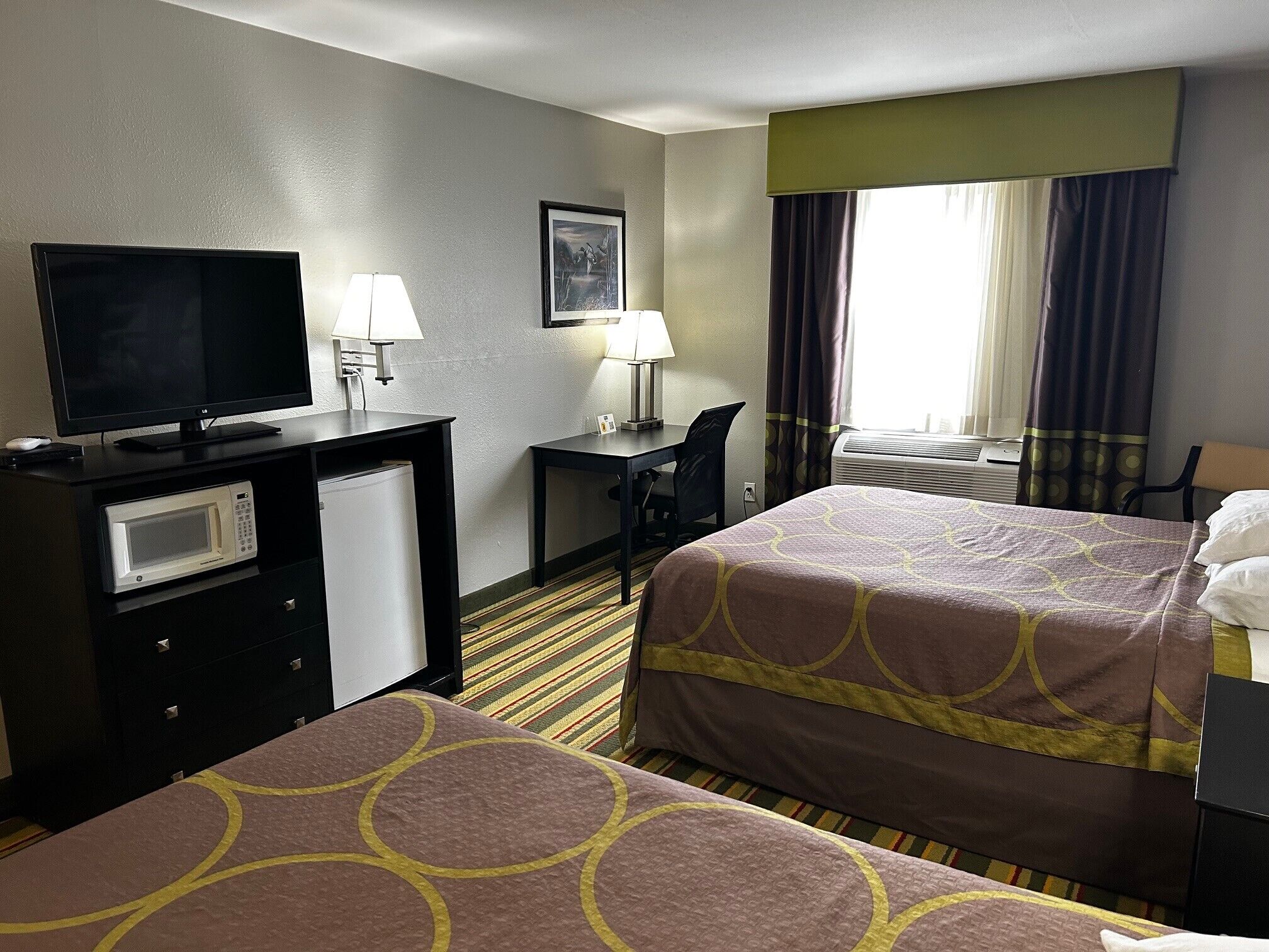 A hotel room with two beds, a desk, a television, a microwave, and a window with curtains.