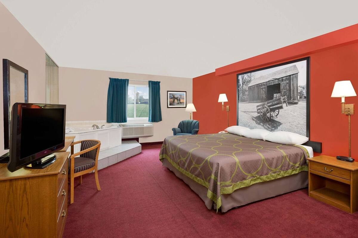 Hotelkamer met een bed, een televisie, een bureau met stoel, een raam met gordijnen, een wandlamp en een schilderij.