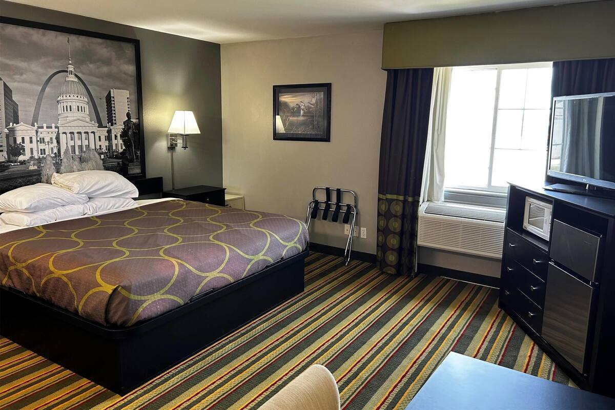 Een hotelkamer met een groot bed, een bureau met stoel, een televisie, een raam met gordijnen, een schilderij aan de muur en een lamp.