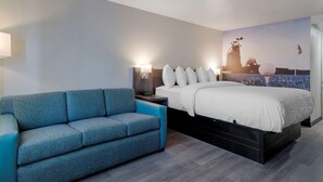 A modern hotel room with a bed, a blue sofa, a nightstand with a lamp, and a wall mural. - Clarion Pointe McDonough (McDonough)