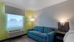 A hotel room with a sofa, a bedside table with a lamp, a nightstand with a clock, and a window with an air conditioner. - Clarion Pointe McDonough (McDonough)