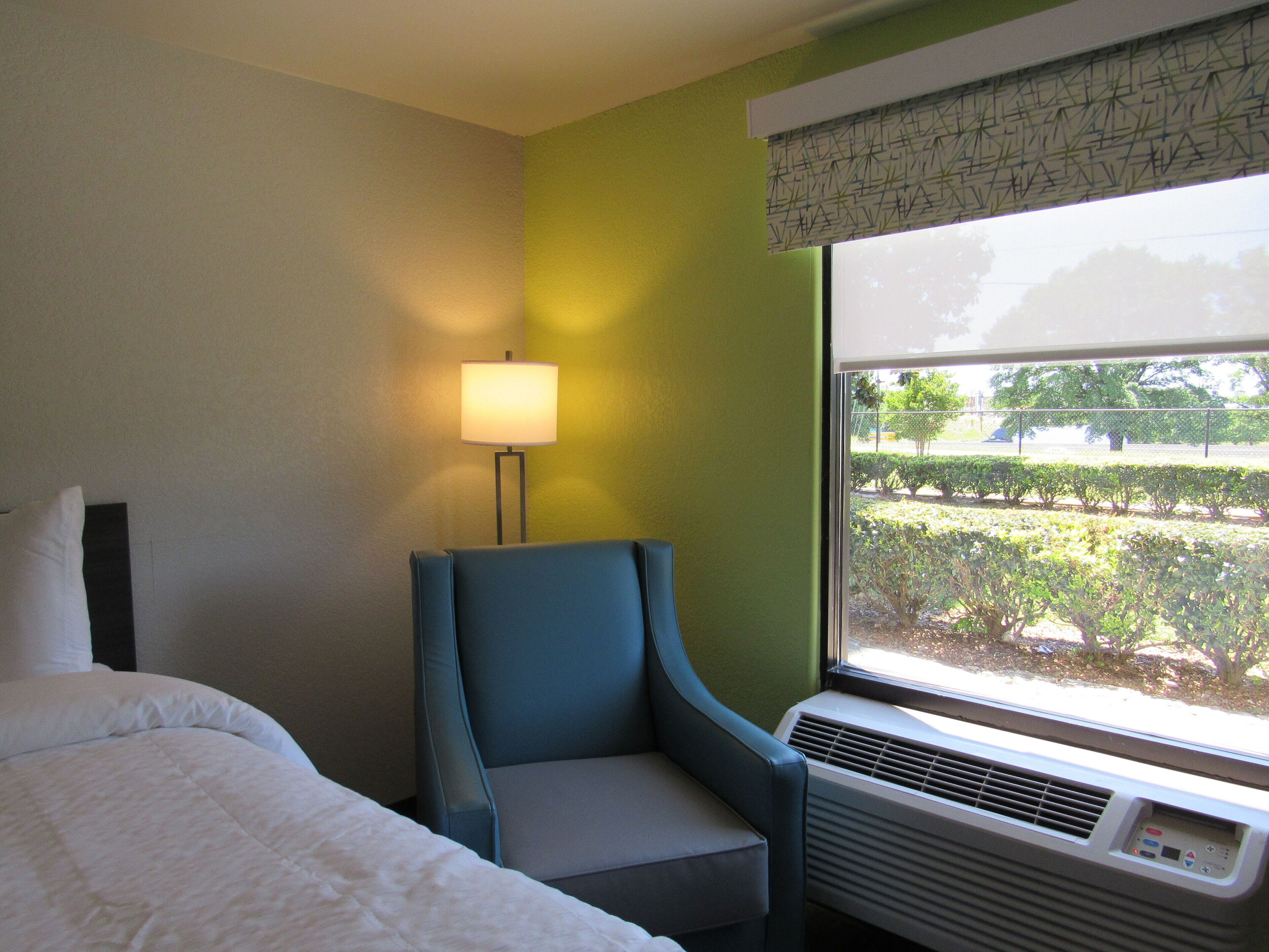 A hotel room with a bed, a chair, a window with a view of a garden, and an air conditioning unit.