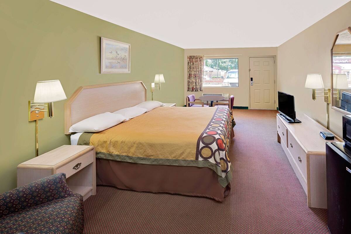 Ein Hotelzimmer mit Bett, Nachttischen, einem Schreibtisch, einem Sessel sowie Fernseher und einem fensterbepflanzten Fenster.