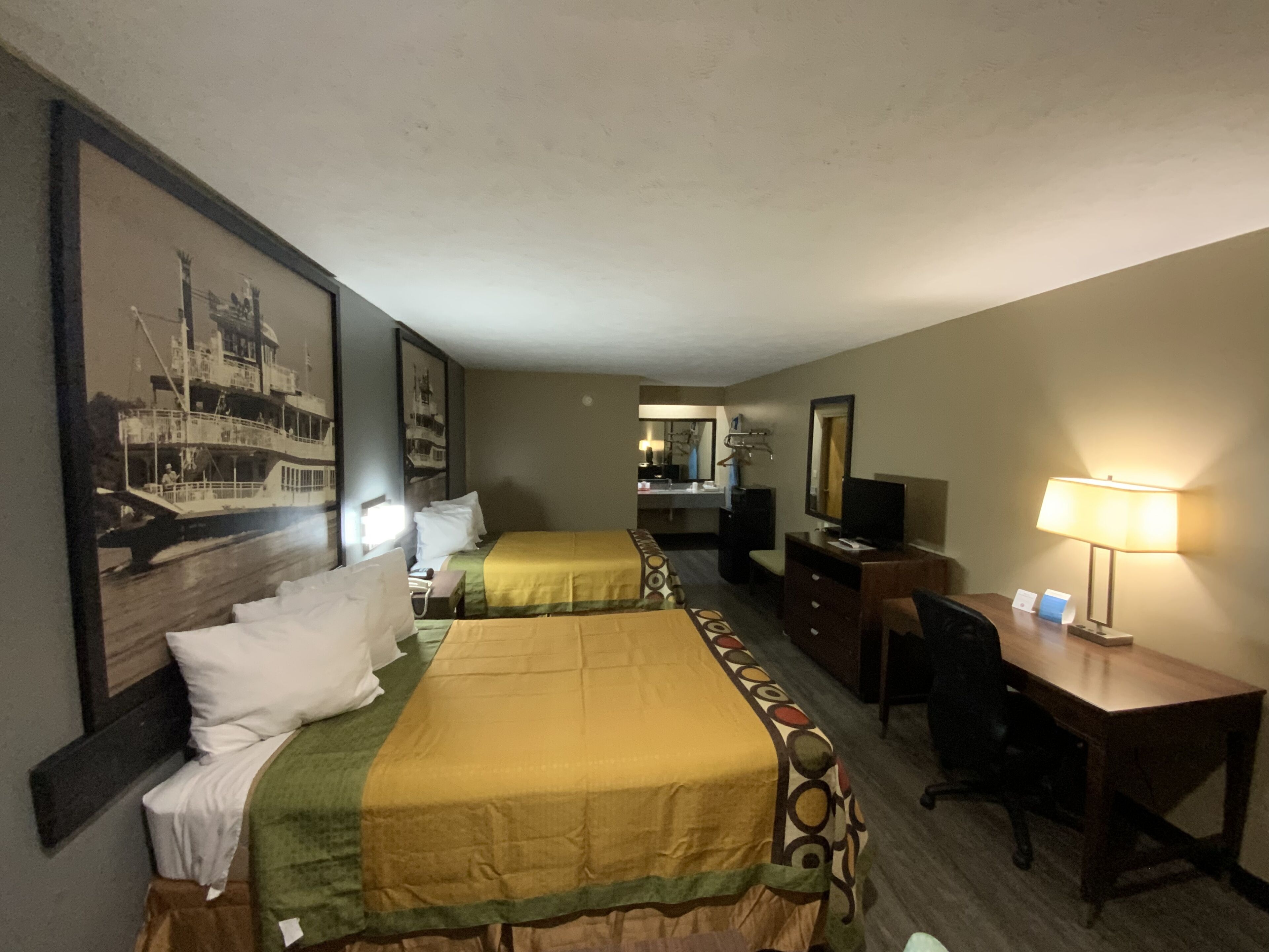 A hotel room with two beds, a desk, a chair, a desk lamp, a television, and a large framed picture on the wall.