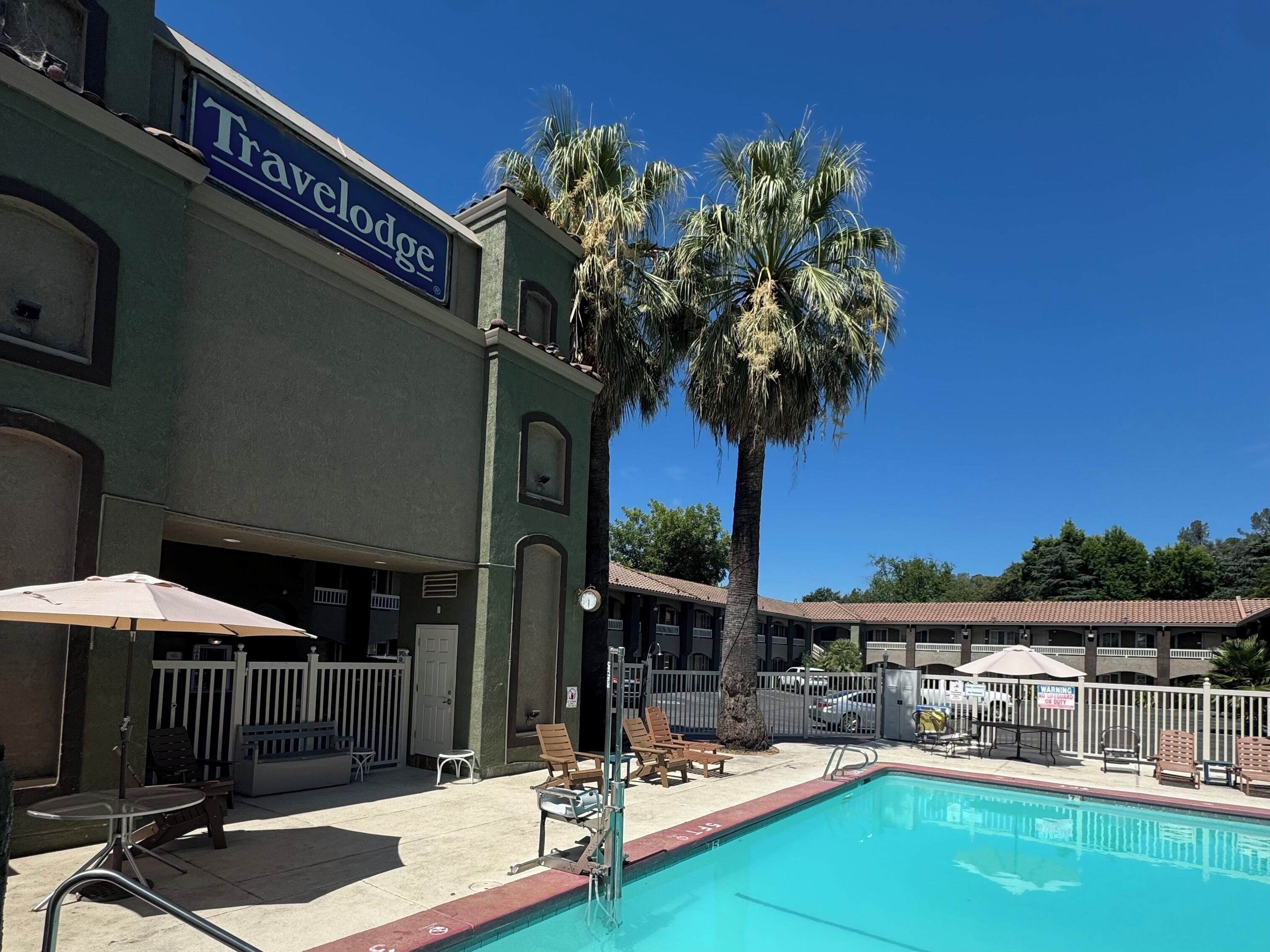 A pool area with lounge chairs and umbrellas, a sign for 'Travelodge,' and a fenced property.