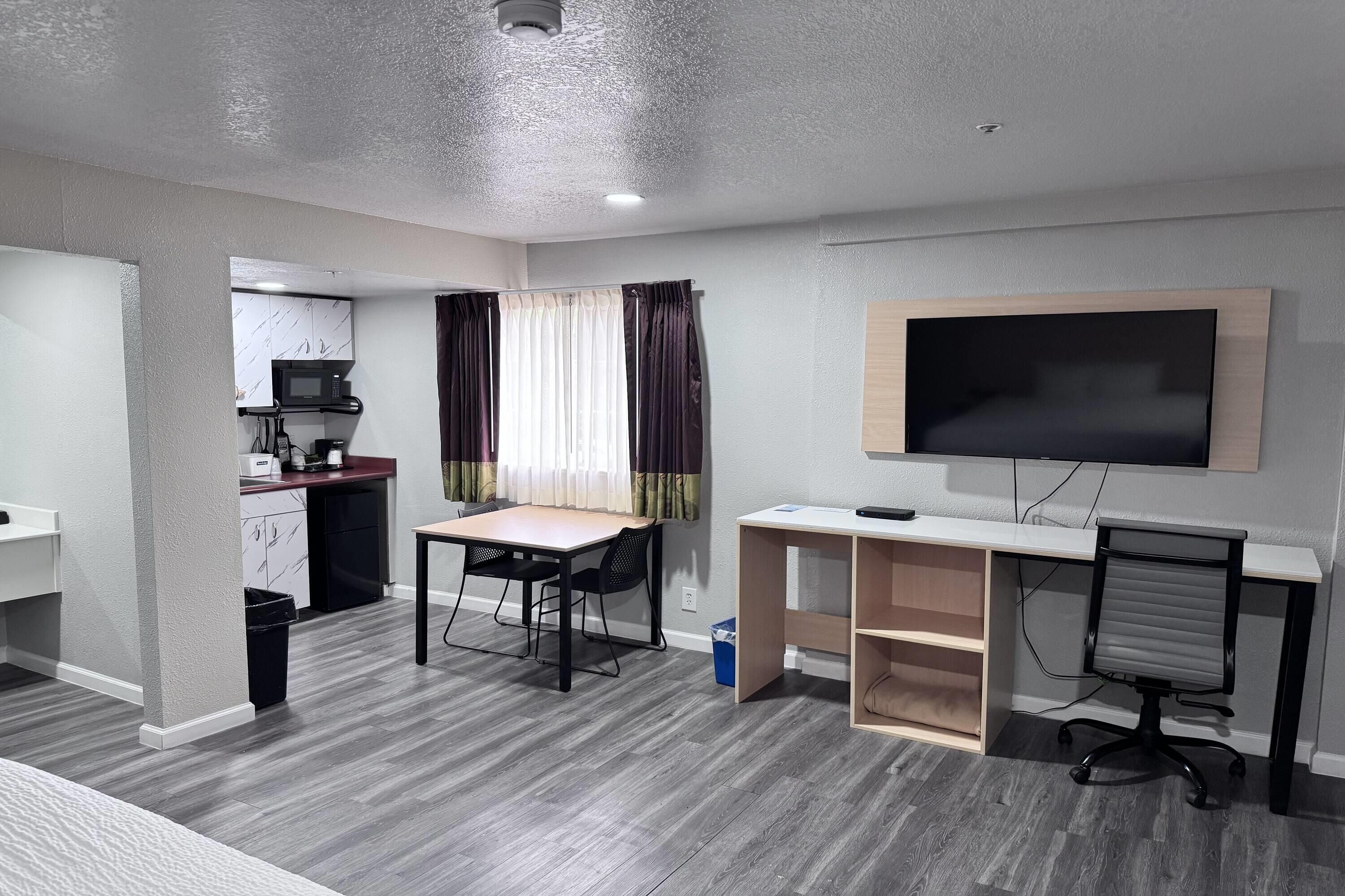 A modern room with a dining table, a TV mounted on the wall, and a kitchenette in the background.