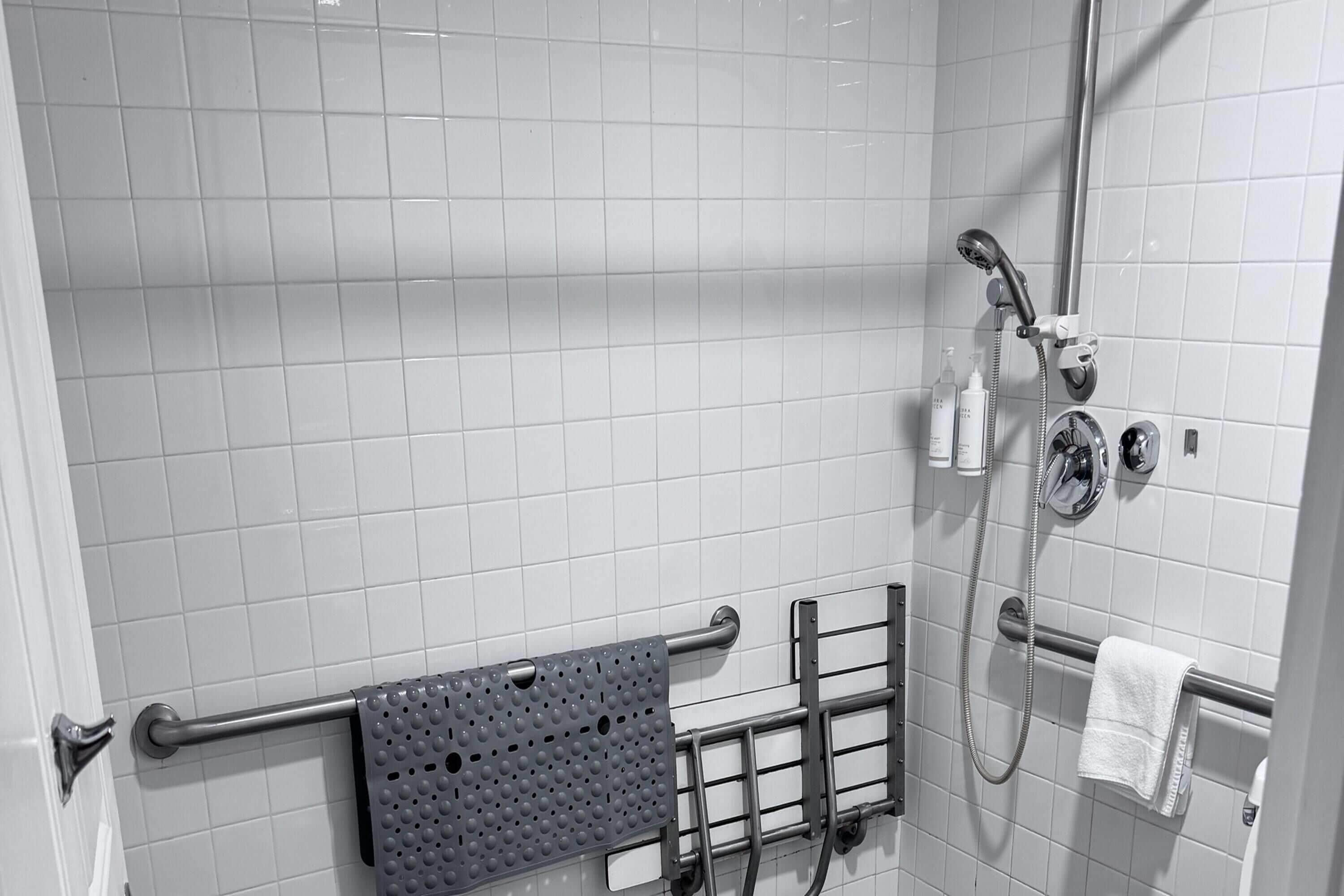 A bathroom with white tiled walls, a shower with a handheld showerhead, a towel rack, and a grab bar.