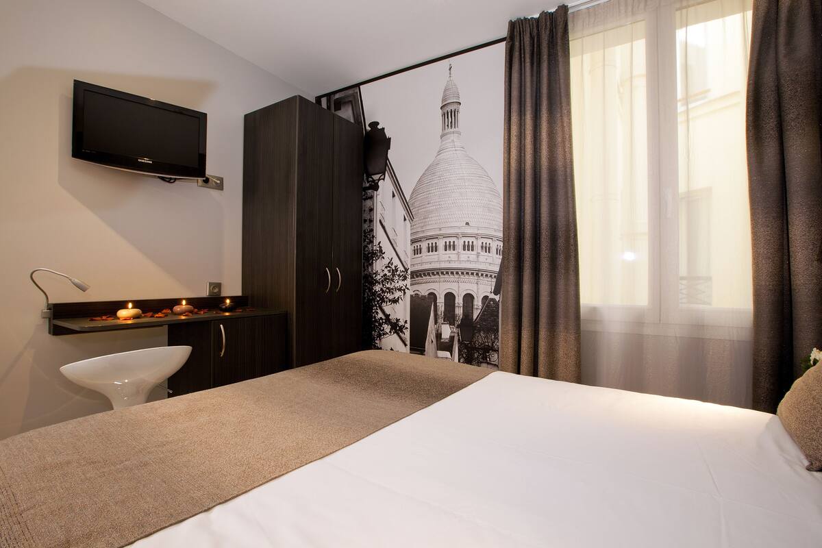 Une chambre d’hôtel avec un lit, un téléviseur fixé au mur, un bureau avec une lampe et une vue sur un immeuble à travers des rideaux transparents.