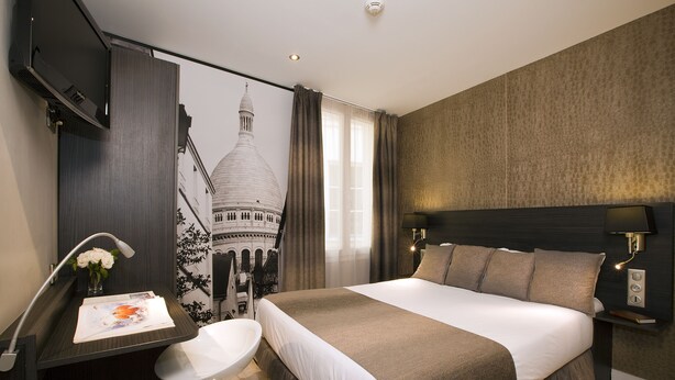 A modern hotel room with a large bed, a desk with a chair, a small table with a vase of flowers, and a wall-mounted TV. - Hotel Eden Opéra (Paris)