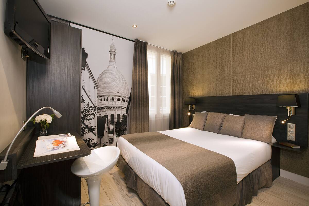 Une chambre d’hôtel moderne avec un grand lit, un bureau avec une chaise, une petite table agrémentée d’un vase de fleurs et un téléviseur fixé au mur.
