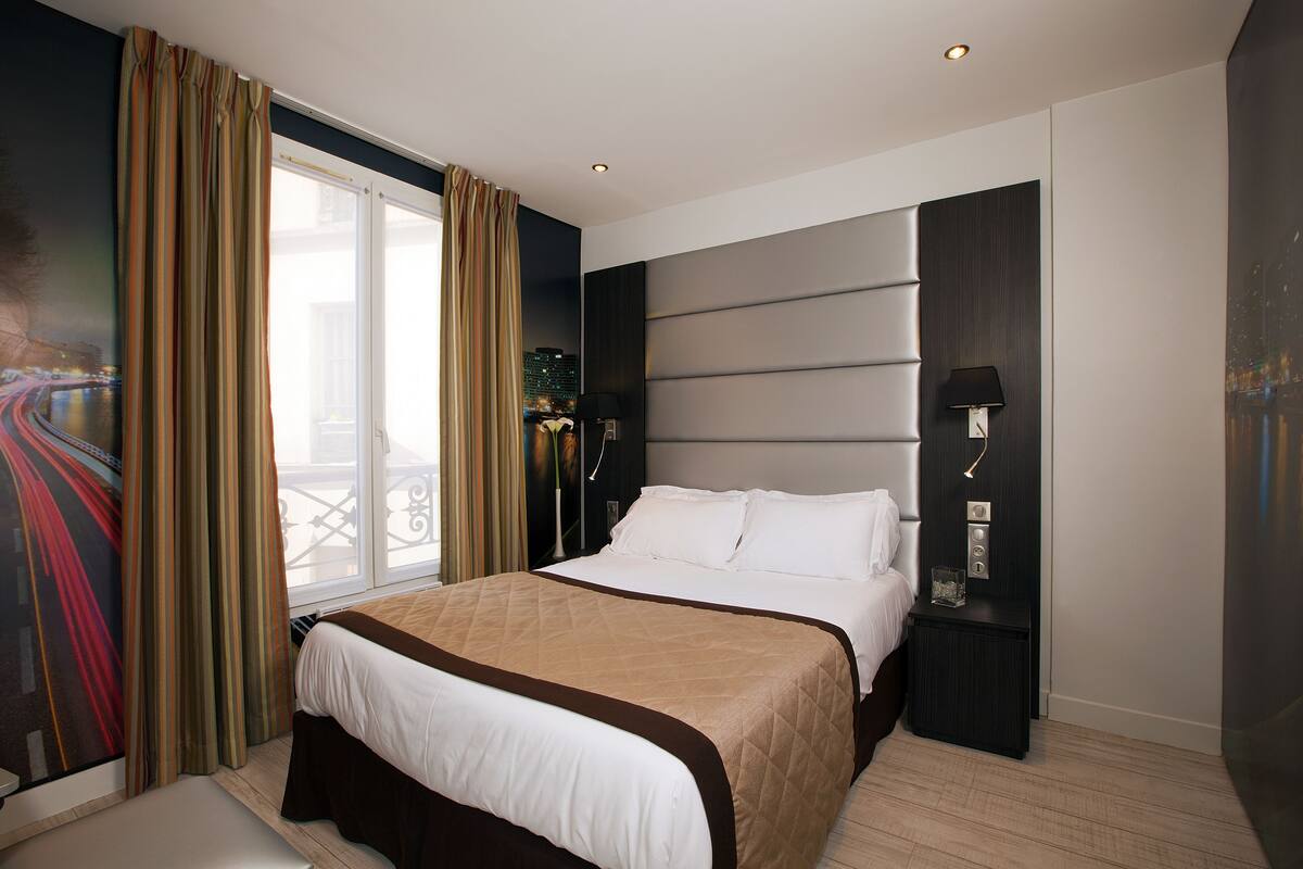 Une chambre d’hôtel moderne, dotée d’un grand lit, d’une table de chevet, d’une lampe et offrant une vue sur la ville par la fenêtre.