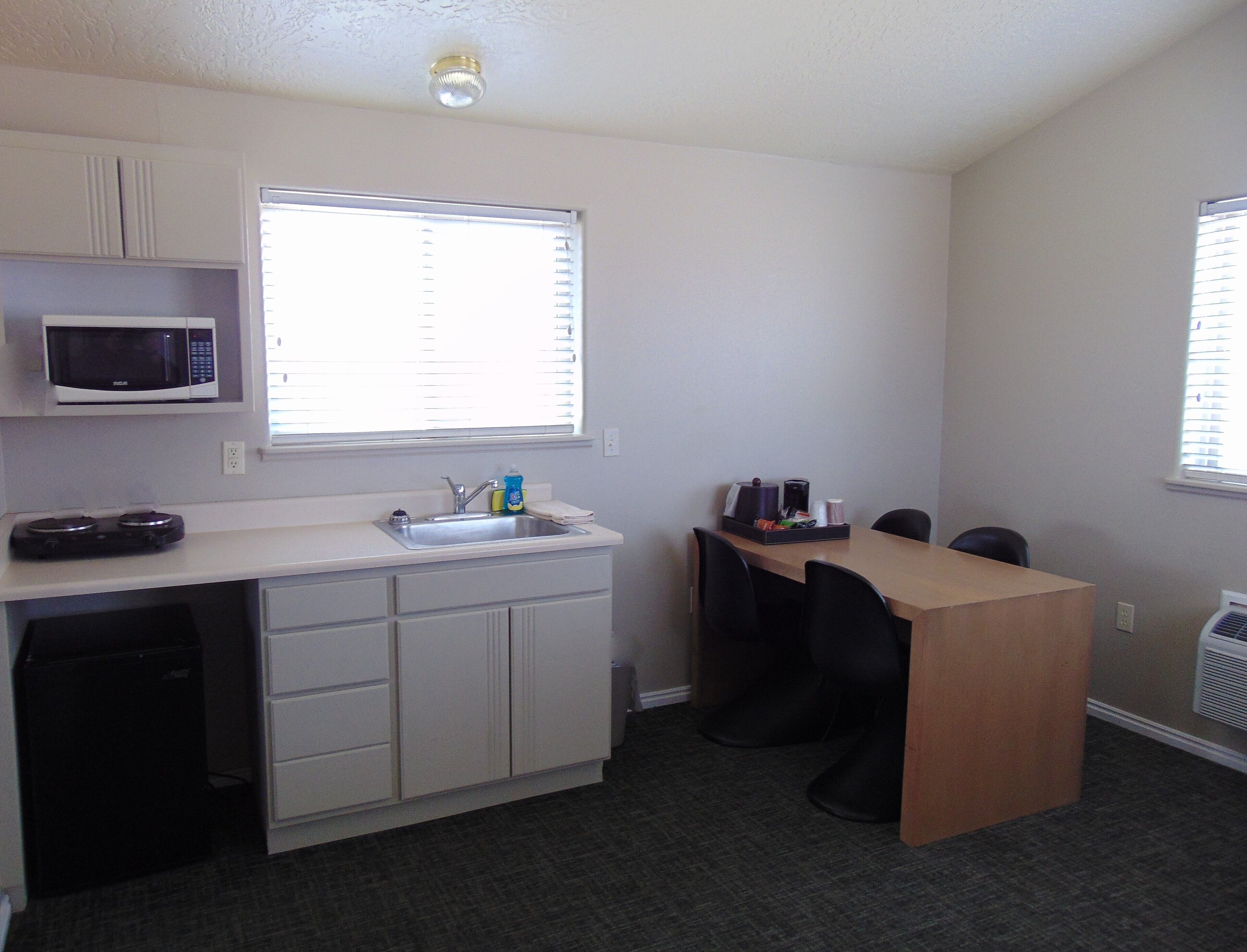 A compact kitchenette with a microwave, sink, and small countertop. There is a window with blinds, a chair, and a table with a tray of items.