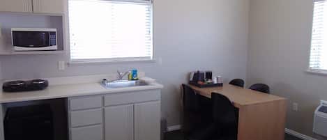 A compact kitchenette with a microwave, sink, and small countertop. There is a window with blinds, a chair, and a table with a tray of items.