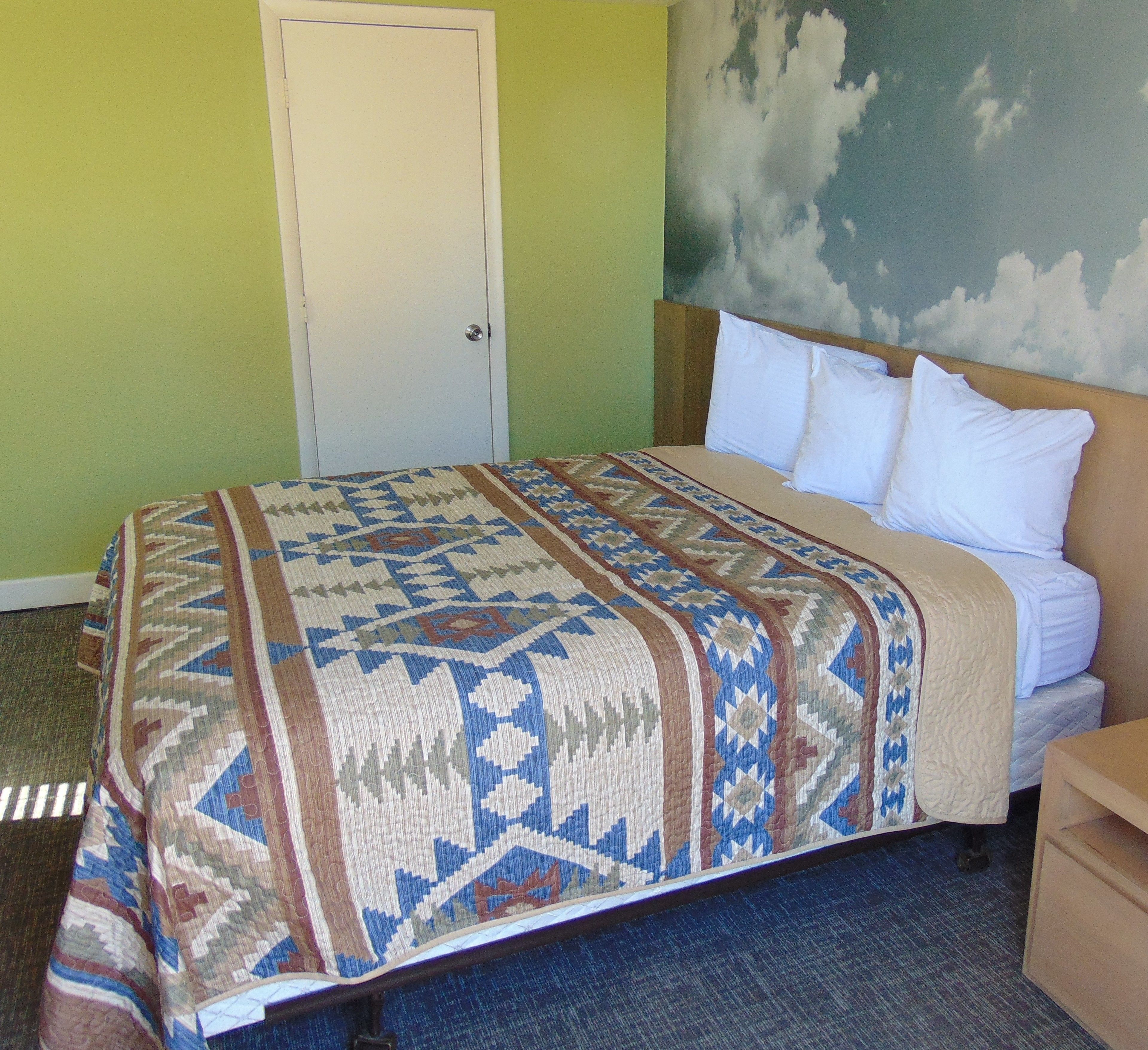 Une chambre d’hôtel avec un lit, une table de chevet, une porte et un mur orné d’une fresque représentant des nuages.