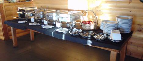 A buffet setup with various food containers, plates, and utensils on a table in a wooden room.