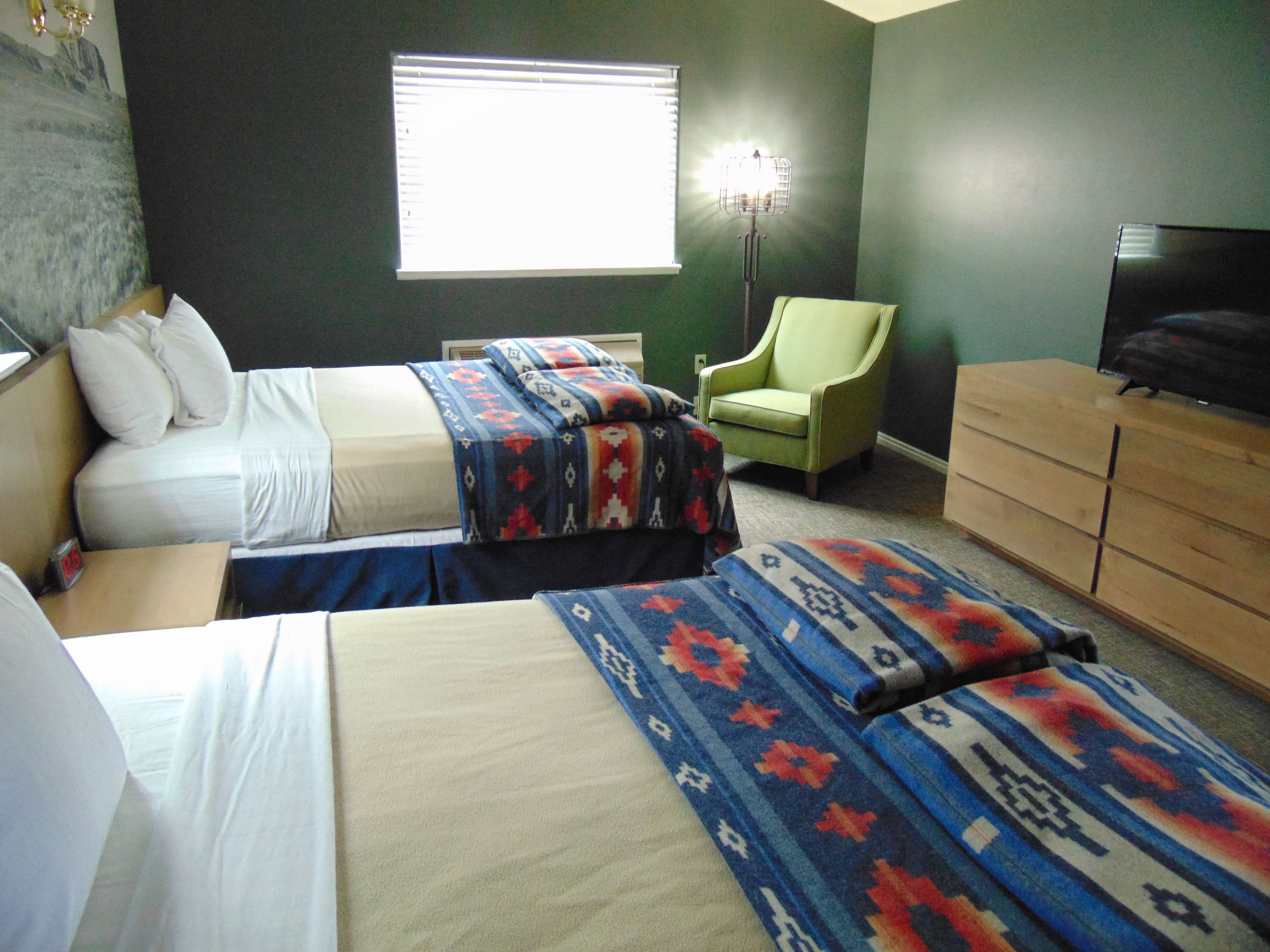 A hotel room with two beds, a green armchair, a wooden dresser, and a television.