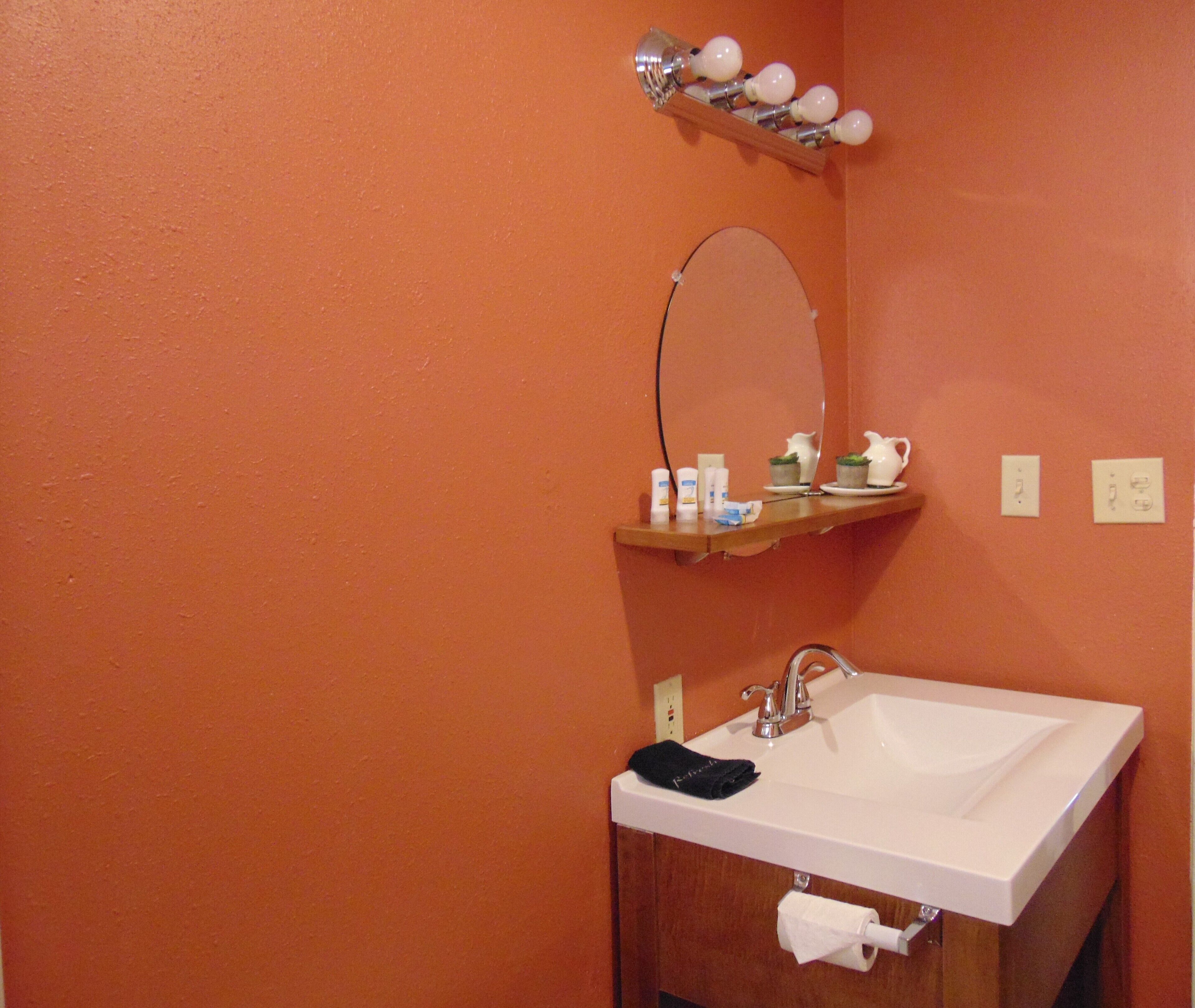 Ein Badezimmer mit orangefarbener Wand, einem weißen Waschbecken, einem Spiegel, einem Ablagefach mit Pflegeprodukten und einem Handtuchspender.
