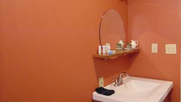Ein Badezimmer mit orangefarbener Wand, einem weißen Waschbecken, einem Spiegel, einem Ablagefach mit Pflegeprodukten und einem Handtuchspender.
