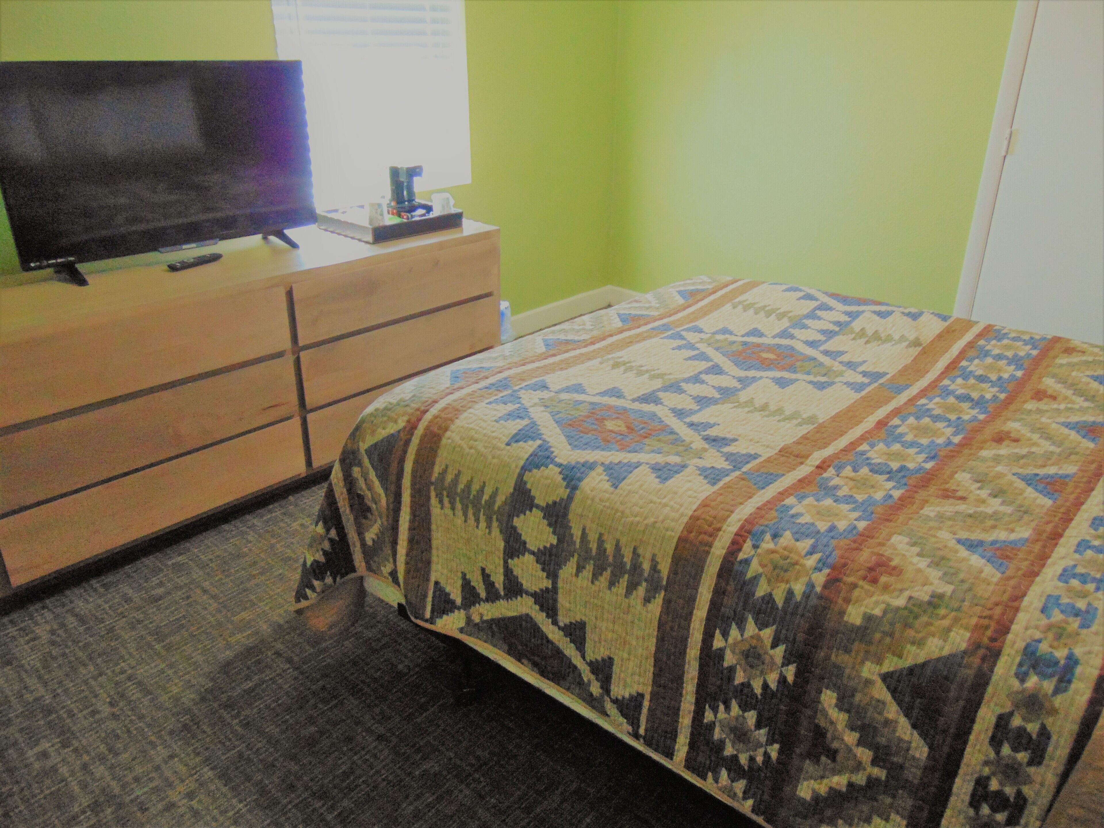 A room with a bed, a dresser, a television on top of the dresser, and a window with blinds.