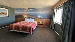 Habitación de hotel con cama, mesita de noche, lámpara, ventana con cortinas y un mural de un paisaje montañoso.