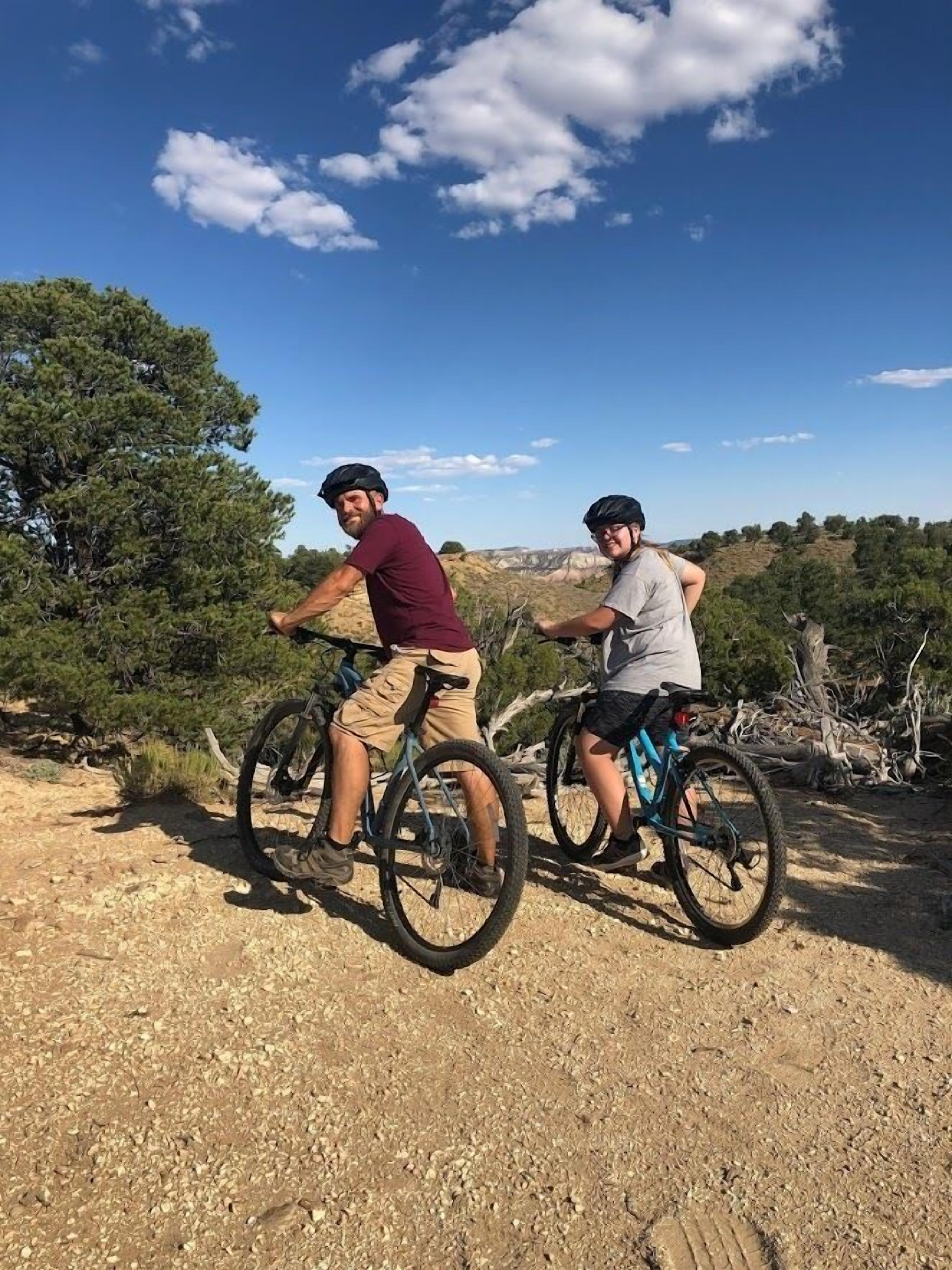 Two people riding bicycles on a dirt path with trees and a clear sky in the background.