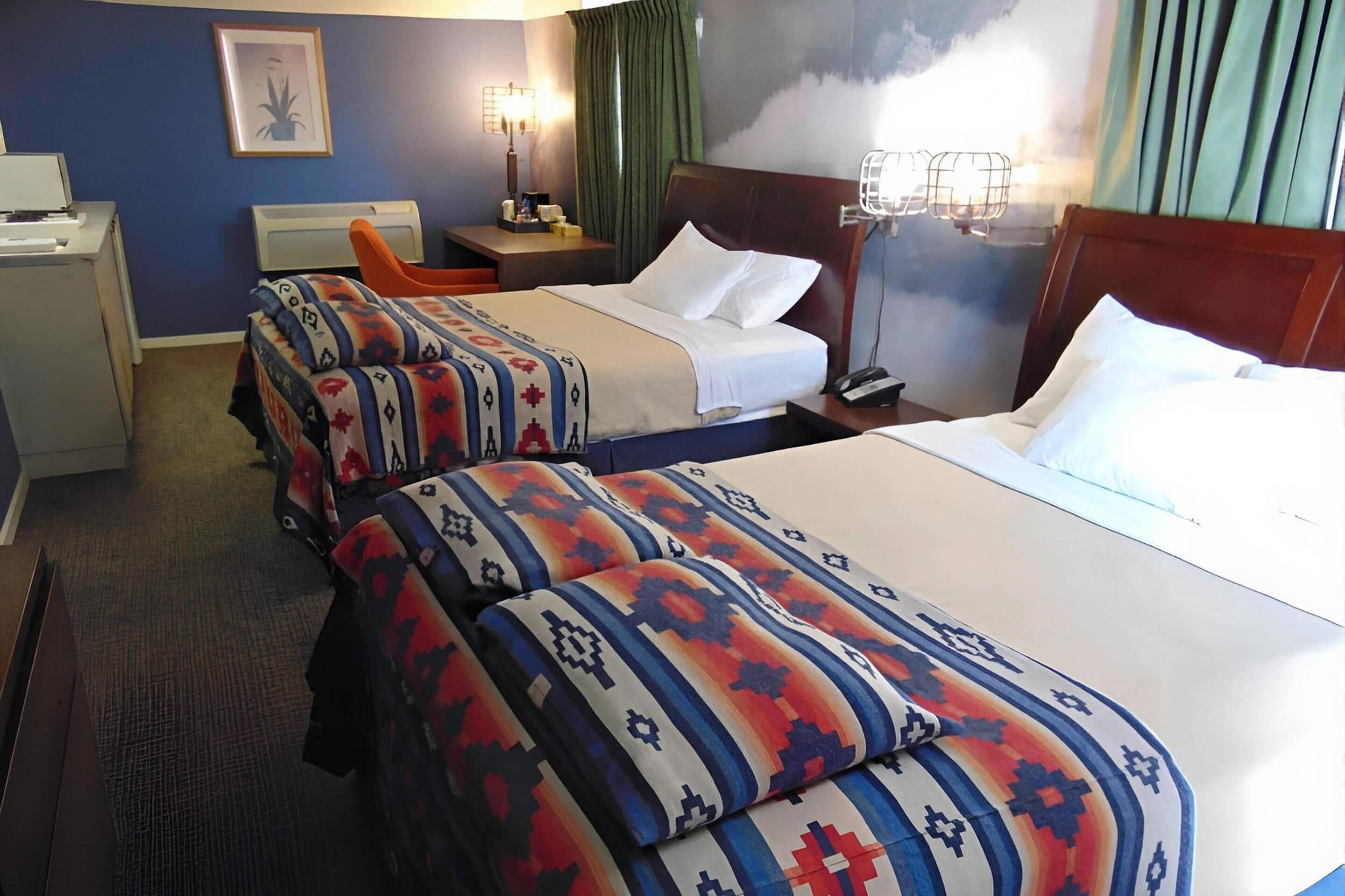 A hotel room with two beds, a desk, a chair, and a nightstand with a lamp.