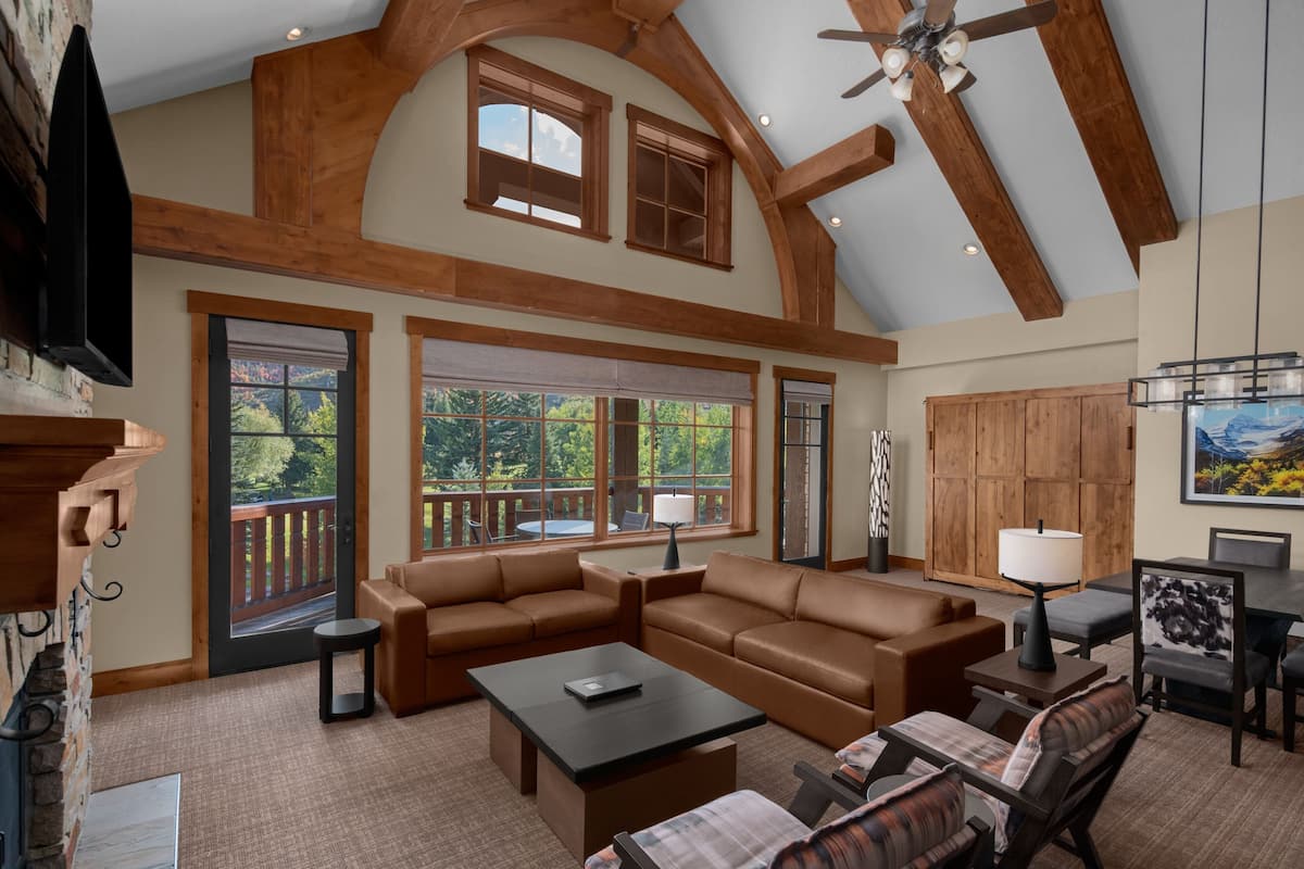 A spacious living room with a large sectional sofa, a coffee table, and a fireplace. There are large windows providing a view of the outdoors.