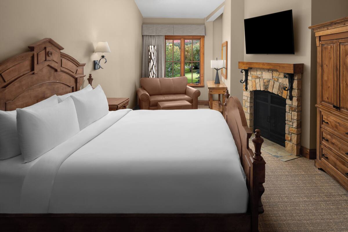 Una cama grande con ropa de cama blanca, cabecera de madera, un sillón marrón, chimenea de piedra y un televisor montado en la pared.