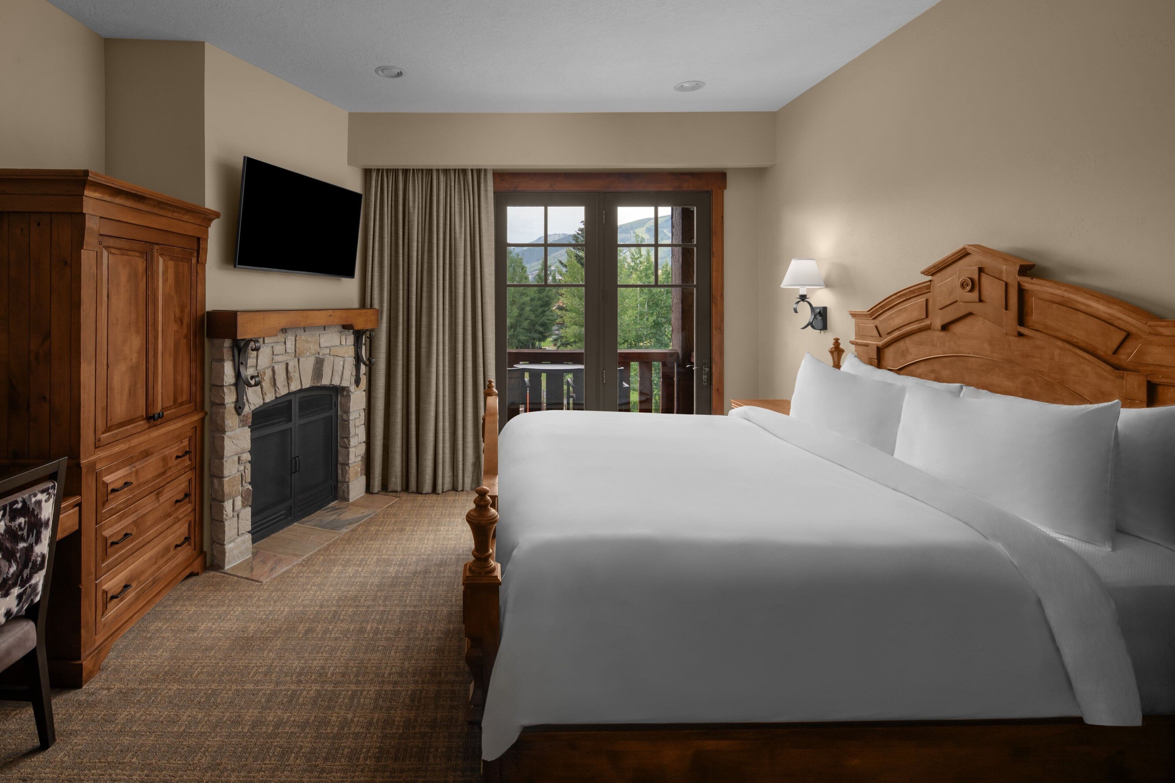 Quarto de hotel com uma cama grande, lareira de pedra, televisão na parede e varanda com vista para o exterior.