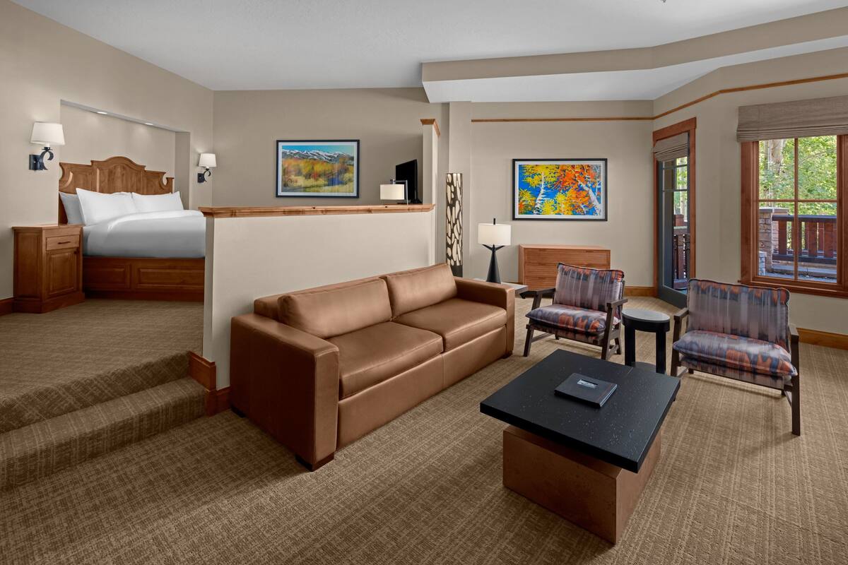 A hotel room with a bed, a brown leather sofa, a small table, two armchairs, a desk, and a window with a view.