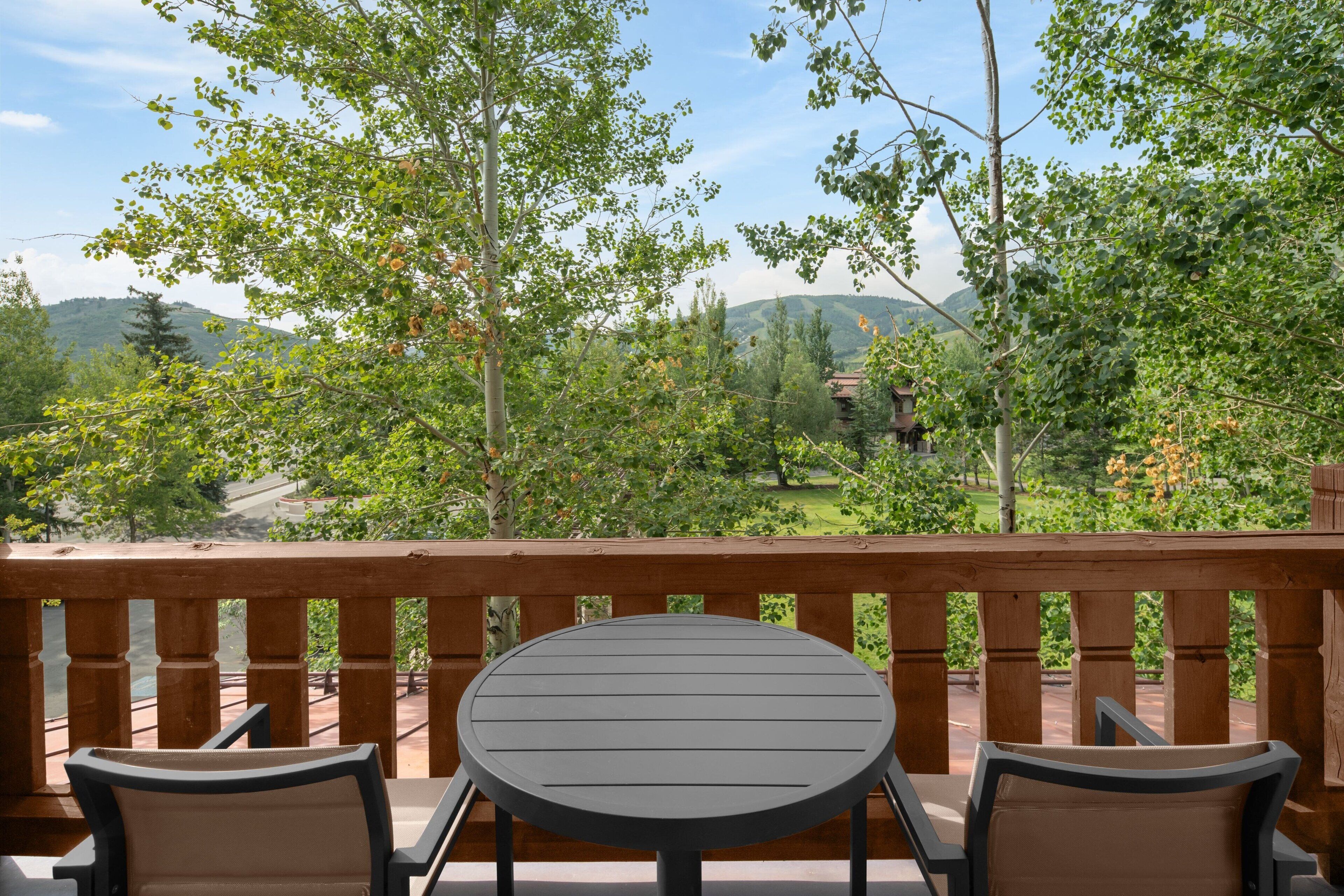 Eine Holzdielen-Terrasse mit einem runden Tisch und zwei Stühlen, mit Blick auf eine malerische Landschaft mit Bäumen und sanften Hügeln in der Ferne.