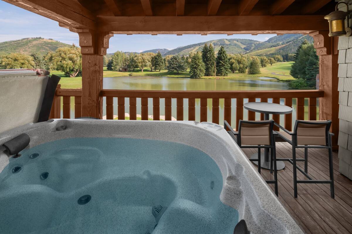 A hot tub with a wooden deck and chairs, overlooking a lake and mountains.