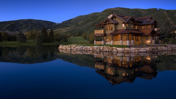 Una casa grande de varios pisos con cimientos de piedra, ubicada cerca de un lago con reflejo claro.
