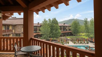 A wooden balcony with a table and chairs, overlooking a pool area with umbrellas and lounge chairs, and a view of buildings and trees.