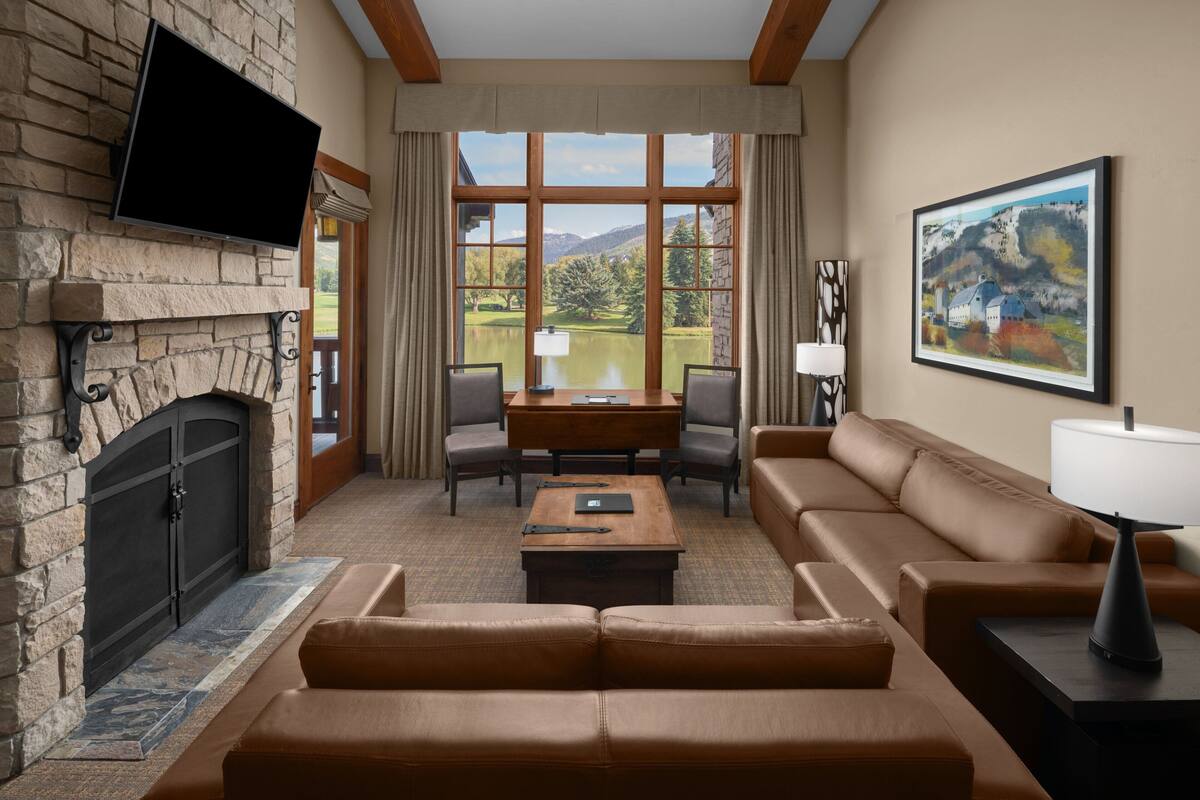 A living room with a fireplace, a television mounted on the wall, a large window with a view of a lake, a sofa, a coffee table, and a painting on the wall.