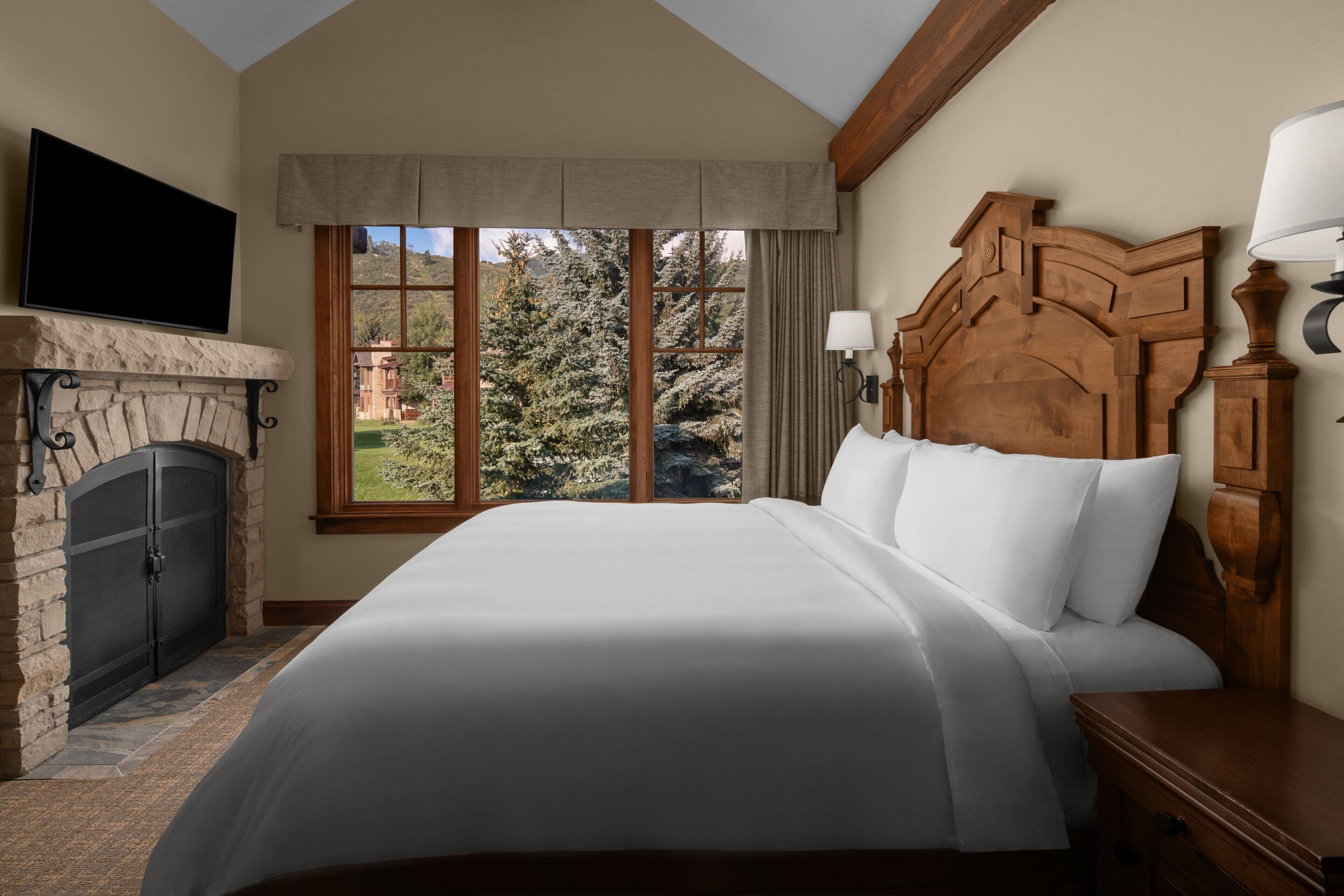 A bedroom with a large bed, a fireplace, a TV mounted above it, and a window with a view of trees.