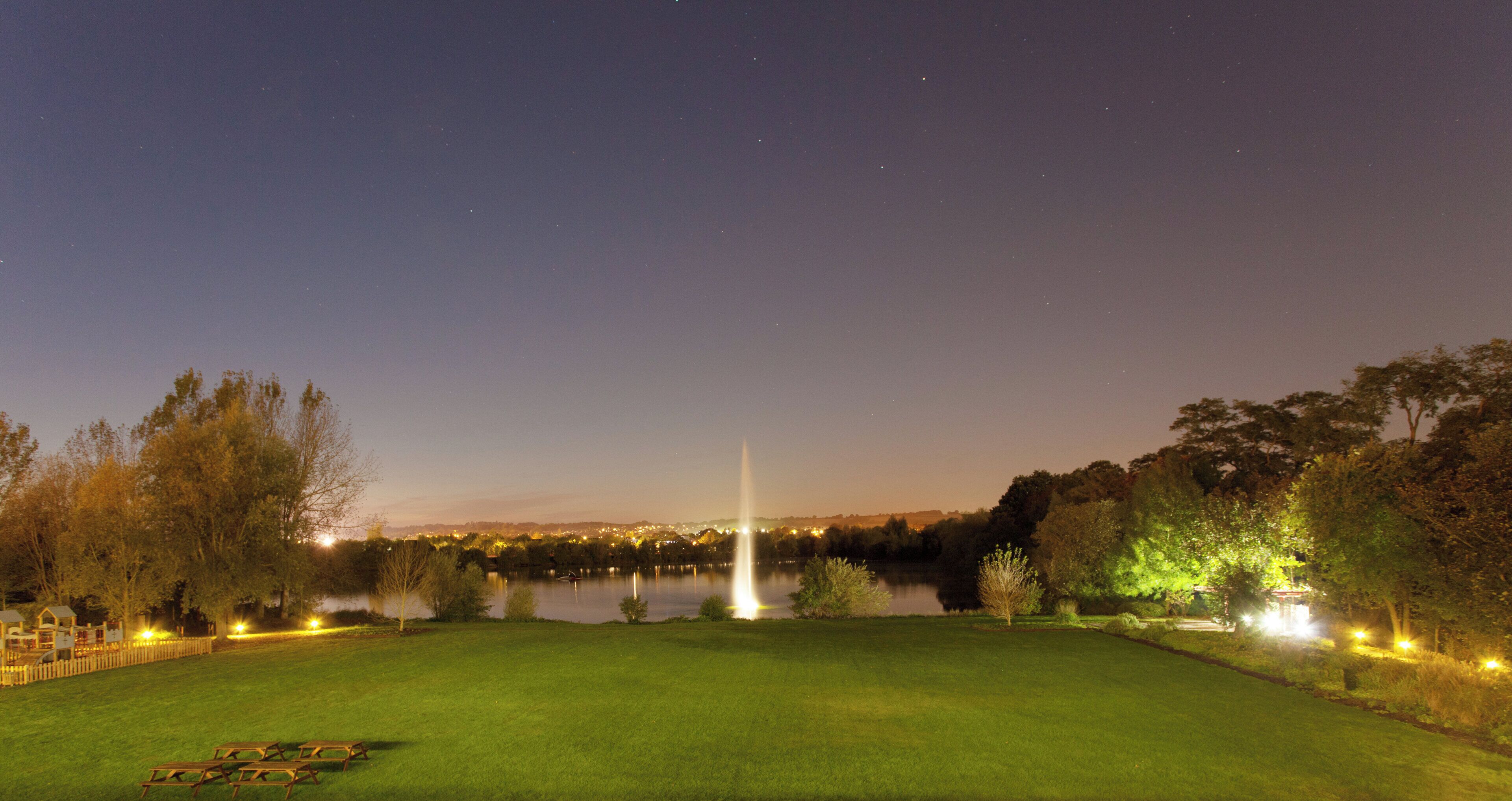 A nighttime view of a park with a fountain, a lake, and a well-lit lawn area.
