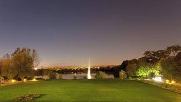A nighttime view of a park with a fountain, a lake, and a well-lit lawn area.
