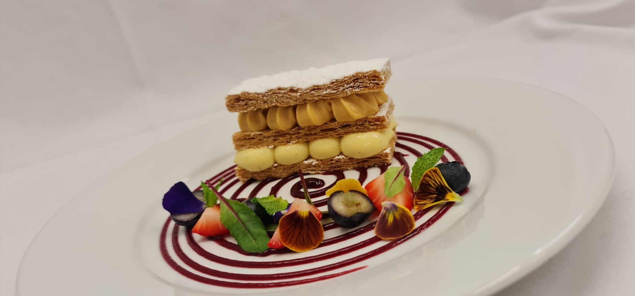 A dessert with multiple layers of cake and cream, garnished with various fruits and leaves on a white plate.