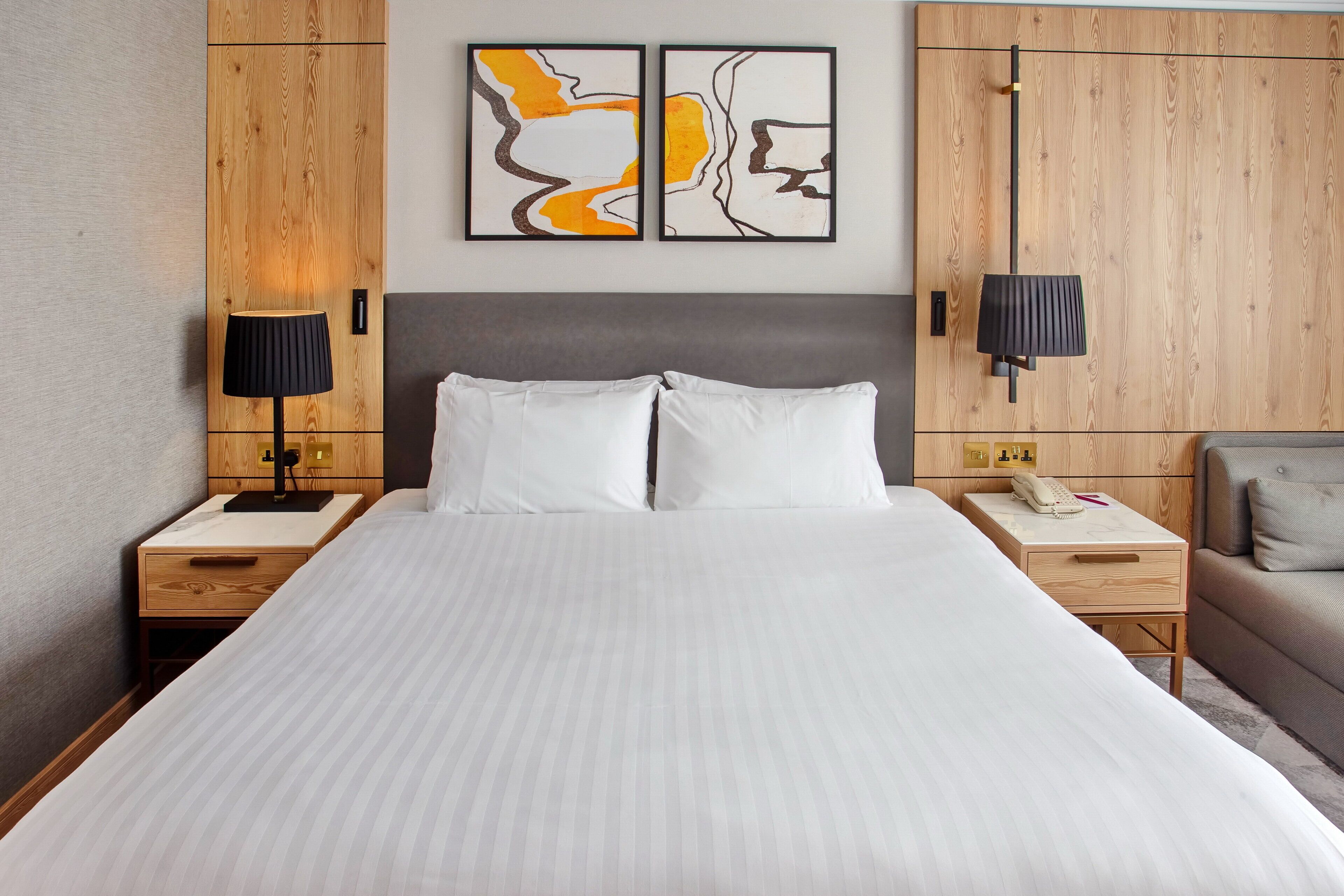 A hotel room with a large bed, two bedside tables, a nightstand, a lamp, a sofa, and two abstract paintings on the wall.