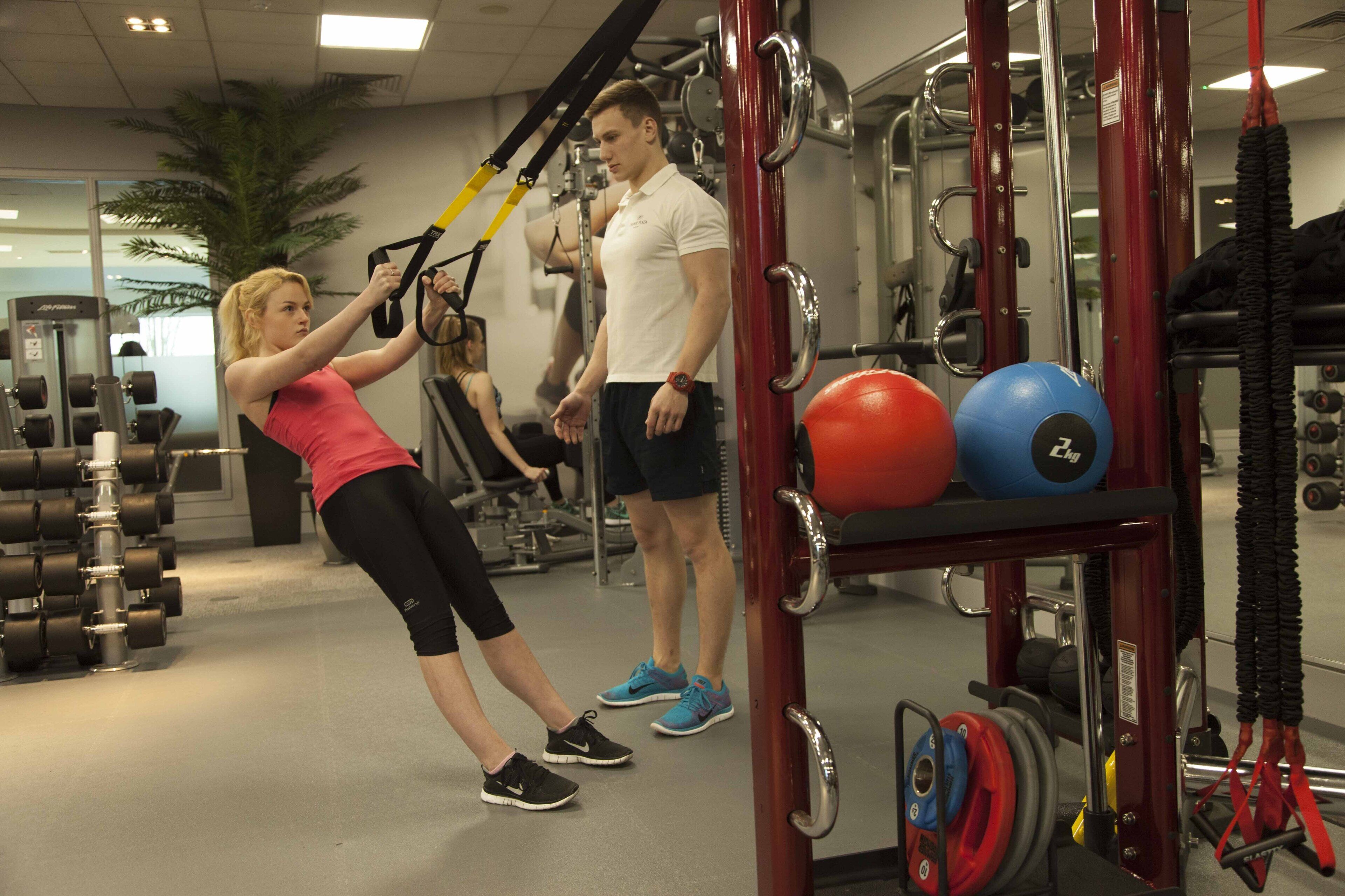 Una palestra con attrezzature per esercizi, tra cui un pallone medicinale rosso e blu, e una persona che utilizza un trainer di sospensione TRX.
