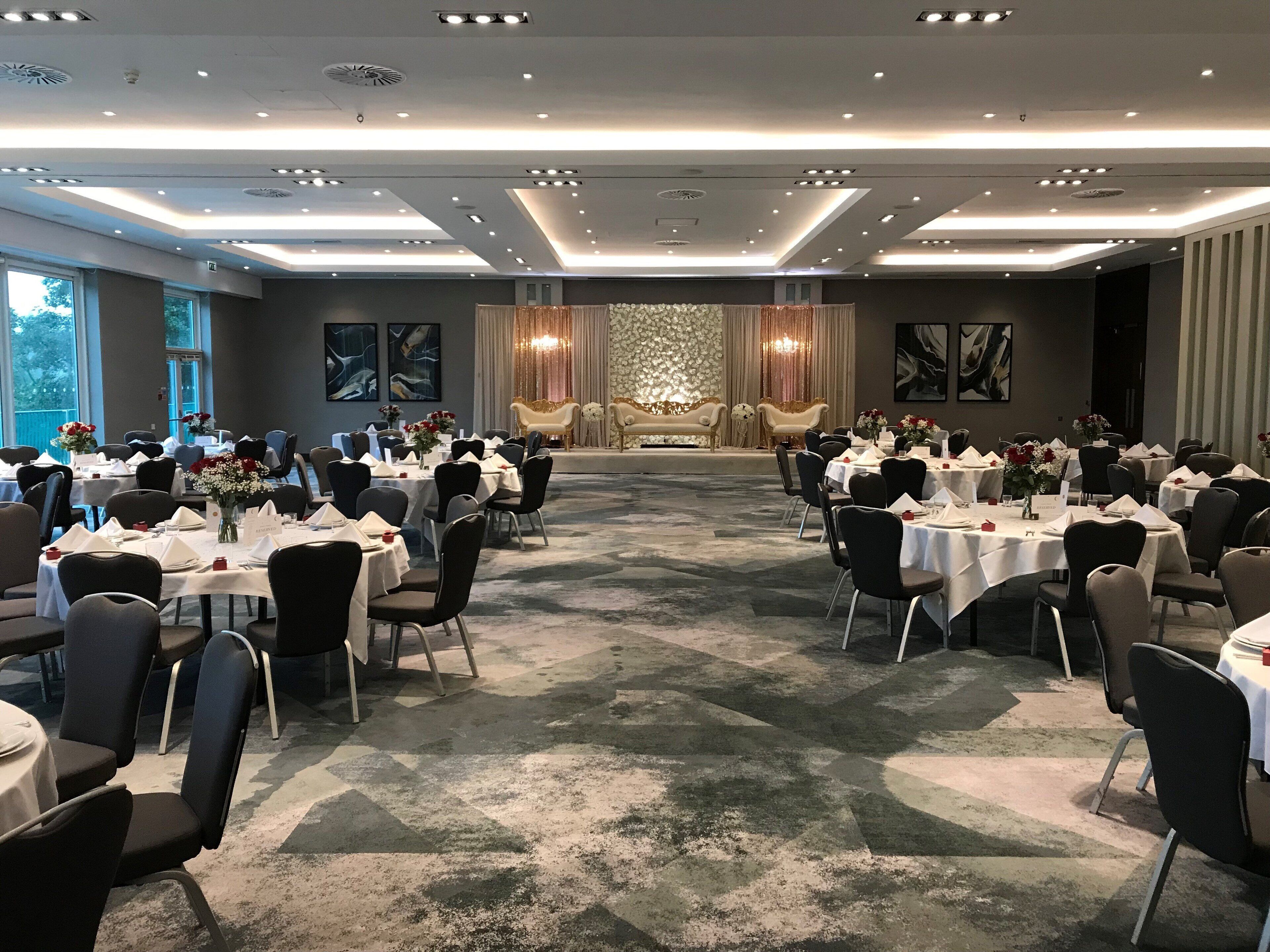 A banquet hall with round tables set for dining, a stage with curtains, and a patterned carpet.