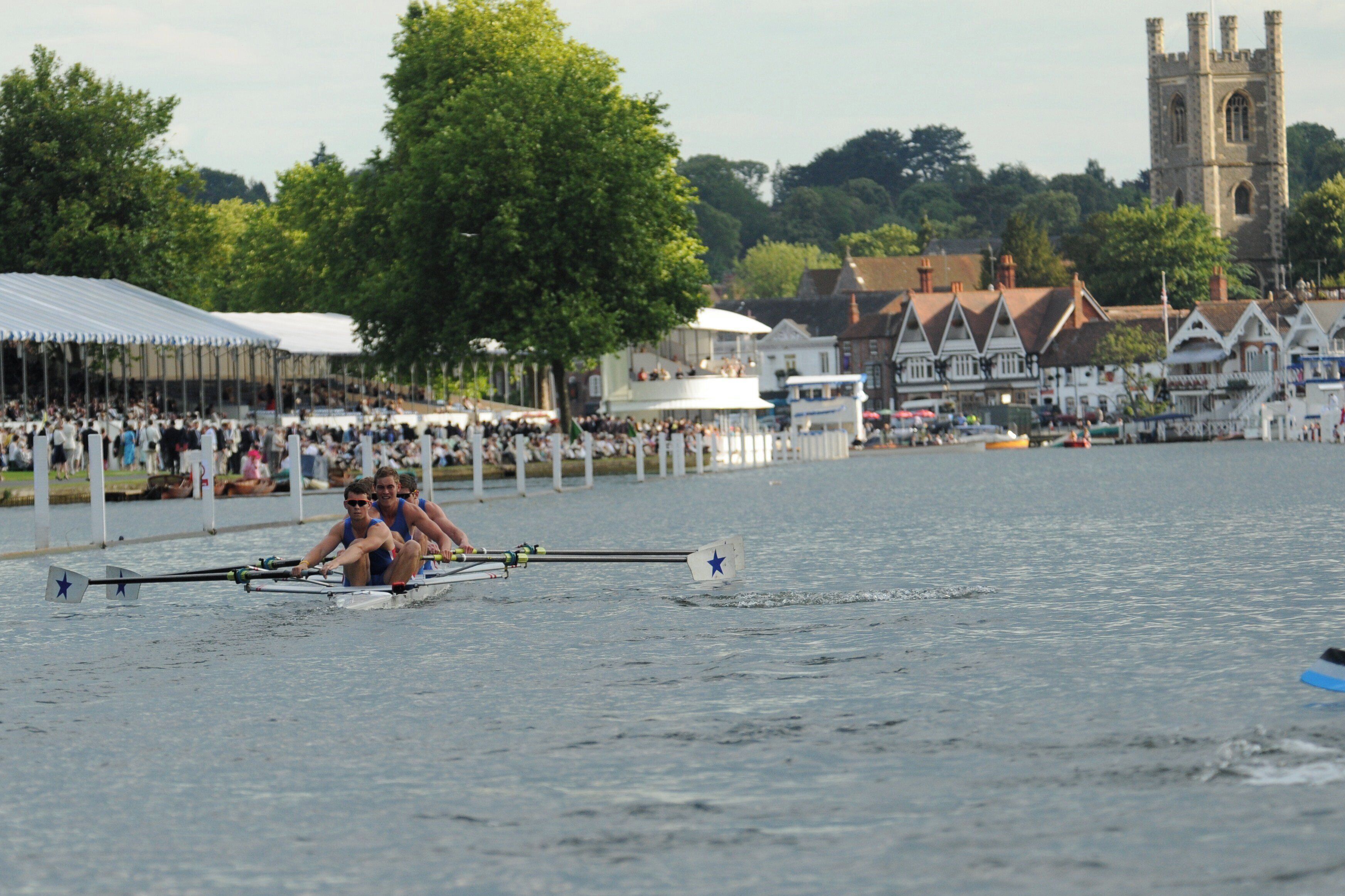 A rowing team in a boat on a river with spectators and buildings in the background.
